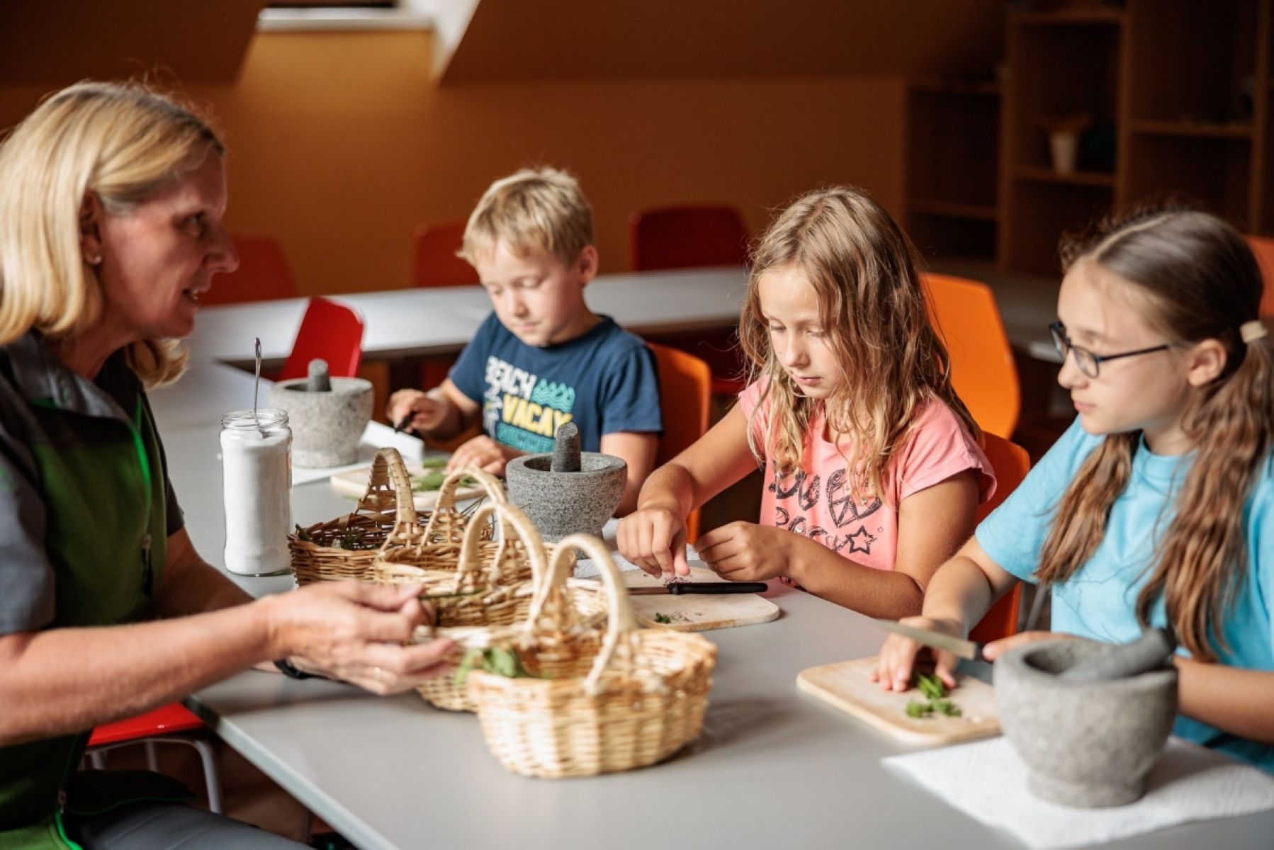 Eine Nationalparkrangerin sitzt 3 Kindern gegenüber. Die Kinder haben Schneidbretter und Kräuter vor sich.